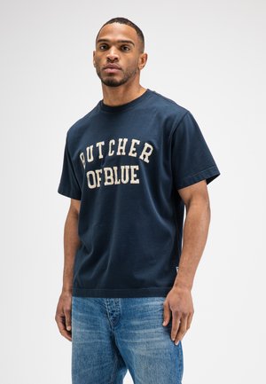 Man wearing navy blue t-shirt with "BUTCHER OF BLUE" text and blue jeans, standing against a plain light background.