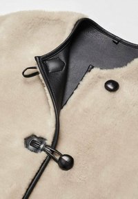 Tissu en shearling beige avec garniture en cuir noir et fermeture à bouton bascule, montrant l'intérieur en cuir lisse et l'extérieur en fourrure texturée.