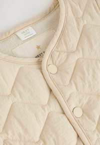 Gepolsterte beige Babyweste mit rundem Ausschnitt, zwei Druckknöpfen und einem weichen Innfutter. Verfügt über ein dezentes Rautenmuster und ein Etikett.