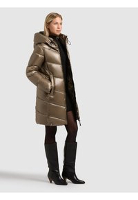 Pufferparka i metallisk olivgrön färg med hög krage, justerbar dragsko i huvan och diagonalt quiltat mönster. Kombinerad med svarta knähöga stövlar.