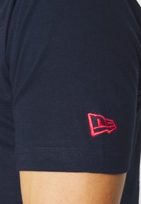 Tmavě modré bavlněné tričko s přiléhavým střihem. Obsahuje červené vyšívané logo na rukávu a hladkou texturu. Jednoduchý kulatý výstřih.