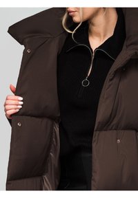 Giacca imbottita marrone con colletto alto, bottoni a pressione e una texture liscia, indossata sopra un maglione nero a coste con scollo con zip.