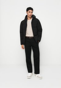 Black puffer jacket with a hood, featuring a quilted design. Worn over a beige sweater, paired with black pants and white shoes.