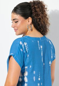 Blå tie-dye topp med korta ärmar, rund halsringning och ett texturerat tyg med vita mönster. Silverfärgade hoops örhängen är synliga.