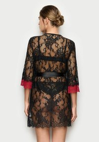 Černý krajkový župan se červenými krajkovými rukávy, vlnkovaným lemem a uvázaným černým páskem. Průhledná textura odhaluje skryté oblečení.