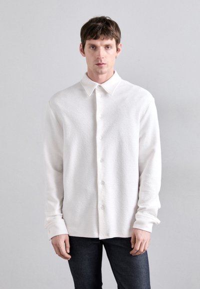 White textured button-up shirt with a collar and long sleeves, paired with dark denim jeans. Simple design, no visible patterns or accents.