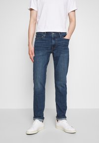 Vaqueros azul oscuro de corte ajustado, fabricados en denim, con cinco bolsillos y un suave desvanecimiento en los muslos. Combinados con zapatillas blancas.