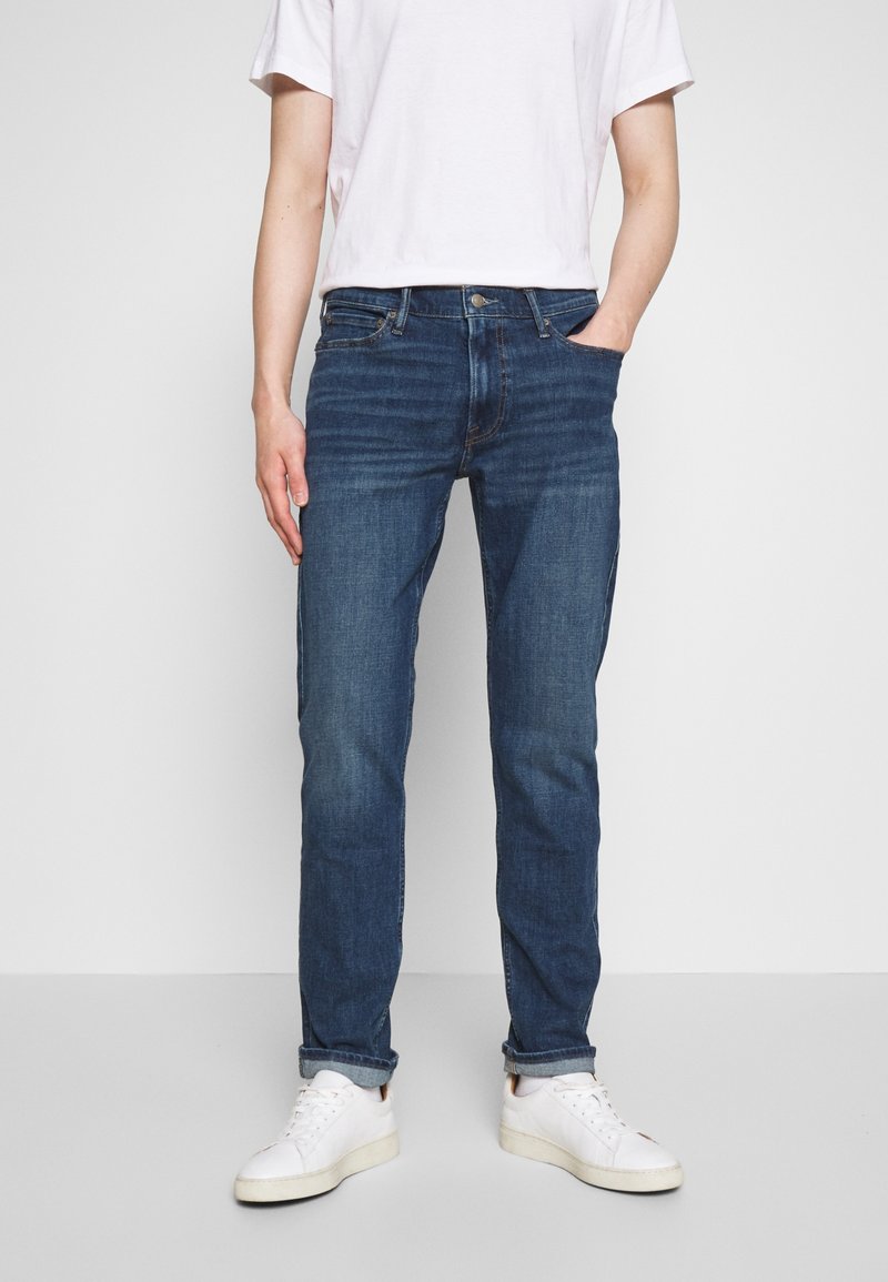 Vaqueros azul oscuro de corte ajustado, fabricados en denim, con cinco bolsillos y un suave desvanecimiento en los muslos. Combinados con zapatillas blancas.