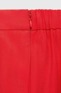 Rode stof met een gladde textuur, voorzien van een ritsluiting en geplooide taille-detail. De stiksels zijn zichtbaar langs de randen.