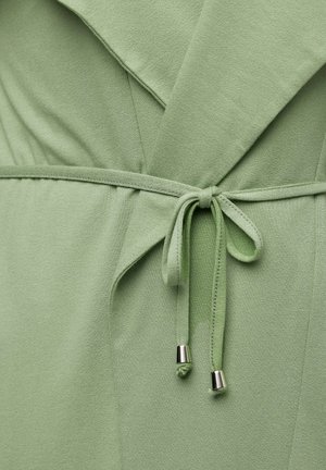 Vihreä kangasvaate, jossa on vyötärön korostus solmittavalla yksityiskohdalla ja metallikoristeilla. Pehmeä rakenne, joka on sileäpintainen ja kerroksellinen.