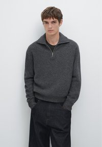 Pull gris côtelé avec un col zippé, associé à un pantalon en denim foncé. Le pull présente une coupe décontractée et un tissu texturé.