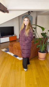 Hosszú lila puffertalpú kabát cipzárral, oldalzsebekkel és kényelmes szabással. Meleg fa padló és beltéri növények előtt.