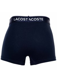 Marineblå bomullshängselbyxor med en vit midja där "LACOSTE" står i blå bokstäver. Enkla, släta texturer och midhög design.