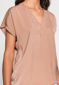 Blusa corta a maniche corte color beige chiaro con scollo a V e vestibilità ampia. Tessuto morbido con un leggero lucido e dettaglio con tasca frontale.