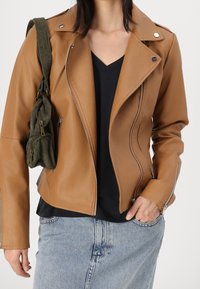 Kobieta w brązowej skórzanej kurtce zapinanej na zamek, nosząca czarną bluzkę w serek, jasnoniebieską spódnicę dżinsową oraz trzymająca oliwkowozieloną torebkę na ramię o teksturowanej powierzchni.