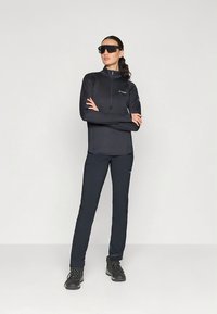 Juodas ilgomis rankovėmis užsegamas viršus, derinamas su juodomis kelnėmis ir žygio batais. Apranga pasižymi tekstūruotu dizainu ir aptempta siluetu.