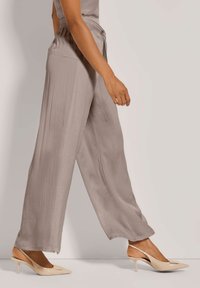 Persoon die loopt in taupe wijduitlopende broek en beige puntige slingback hakken tegen een egale achtergrond.