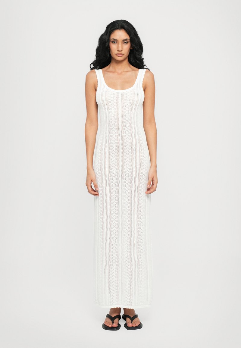 White, knitted maxi dress with a scoop neckline, featuring a textured, lace-like pattern and side slits. Paired with black flip-flops.