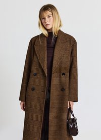 Brauner karierter Doppelreiher-Mantel mit schwarzen Knöpfen, großem Kragen und lockerer Passform. Kombiniert mit einem dunklen Oberteil und einer kleinen dunklen Handtasche.