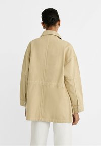 Světle béžová oversized bunda s límečkem, dlouhými rukávy a zapínáním na knoflíky na manžetách. Obsahuje šev, který probíhá vertikálně po zadní straně. Hladký materiál.