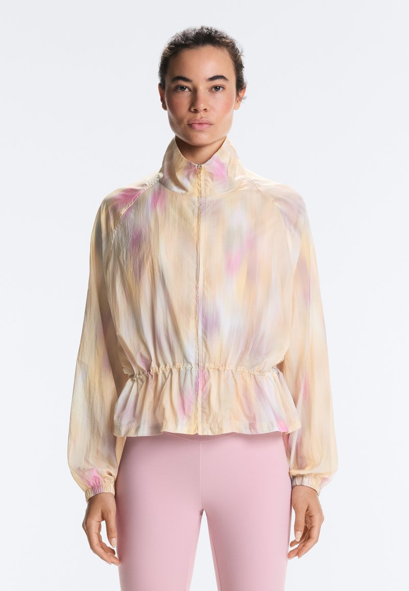 Femme portant une veste zippée de couleur pastel avec taille et poignets élastiques, associée à des leggings rose clair, debout sur un fond blanc.