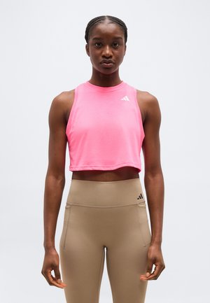Kvinde stående foran, iført en lyserød ærmeløs crop top og højtaljede beige leggings med Adidas-logoer.
