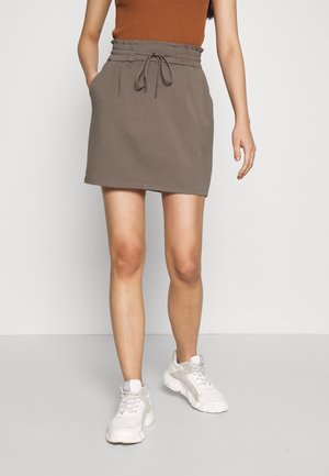 Kobieta w brązowej mini spódnicy ze ściągaczem i kieszeniami oraz białych sneakersach, stojąca na tle jednolitego jasnego tła.