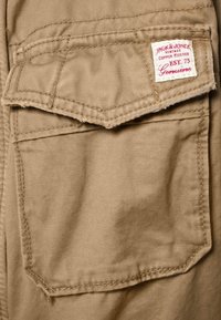 Beżowa kieszeń z tkaniny z detalami przeszyć; posiada metkę z napisem „Jack & Jones” w czerwonym druku, przymocowaną na górze kieszeni.