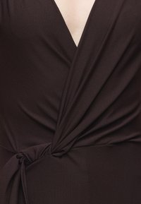 Gros plan d'un tissu marron foncé enroulé et noué à la taille, montrant un décolleté en V et un matériau texturé.