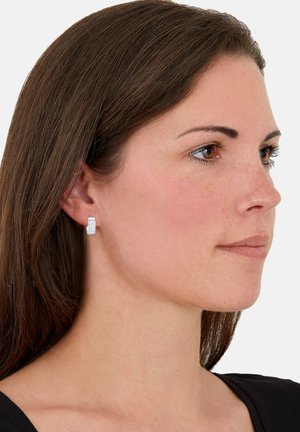 Mujer con cabello castaño lacio que lleva pendientes rectangulares de plata, mirando a la derecha con una expresión neutral.
