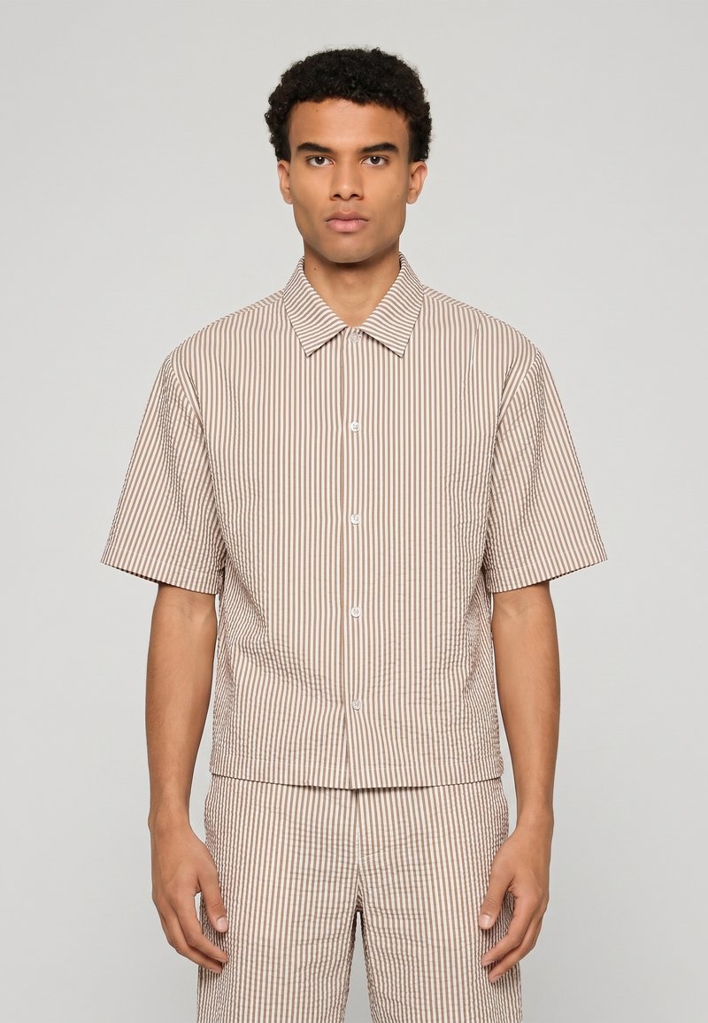 Homme portant une chemise à manches courtes rayée verticale beige et blanche assortie et un pantalon, debout devant un fond uni.