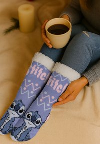Calze di lana viola con polsini bianchi e soffici, caratterizzate da un design di personaggio e motivi geometrici in rosa e blu. Una tazza di caffè in mano.