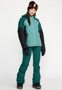 Veste de ski teal et noire avec une fermeture éclair à l'avant, capuche ajustable, associée à un pantalon bleu foncé et des bottes noires, créant un look hivernal coordonné.