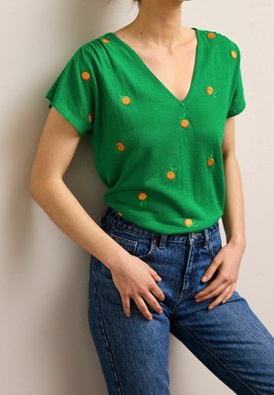 Kvinde iført grøn V-hals kortærmet top med broderede orange frugter og højtaljede blå jeans, står med hænderne i siden.