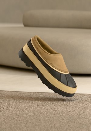 Scarpa slip-on marrone e nera con suola spessa che fluttua sopra un tappeto beige di fronte a un divano beige.