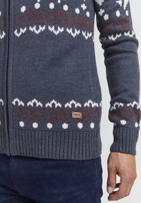Marineblauer Strickpullover mit weißen und braunen Schneeflockenmuster, gerippte Bündchen und Saum, getragen mit dunkelblauen Jeans, die rechte Hand entspannt.