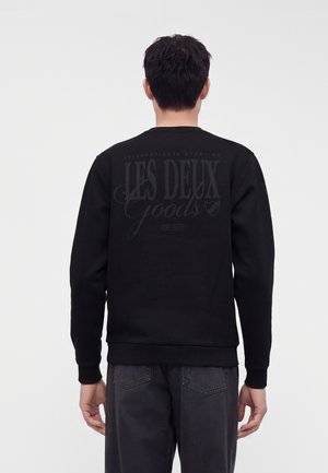 Mand iført sort sweatshirt med "Les Deux Goods" trykt på ryggen, kombineret med mørkegrå jeans, stående mod en hvid baggrund.