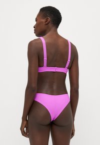 Rožinė bikini komplektas su tekstūruotu audiniu, reguliuojamomis petnešėlėmis ir sagtimi užsegti nugaroje. Dizainas apima žemai sėdintį apatinį trikotažą.