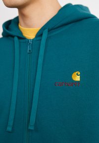 Mėlynos žalią zip-up džemperį su raišteliais; turi siuvinėtą geltoną ir raudoną logotipą; minkšta tekstūra; po džemperiu yra sluoksniuota geltona marškinė.