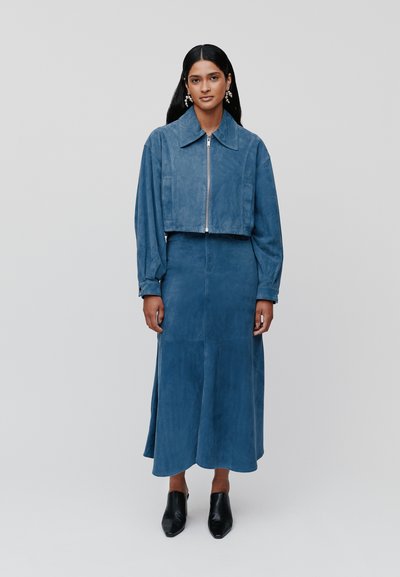 Giacca corta in suede blu con zip frontale e maniche lunghe abbinata a una gonna lunga fino alla caviglia dello stesso colore. Scarpe nere con punta a punta.