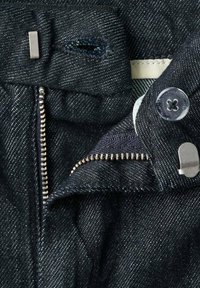 Jeans de mezclilla oscura con cierre de cremallera metálica, cierre de botón y una pequeña etiqueta de tela. La tela presenta una textura sutil y un ligero brillo.