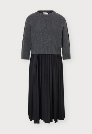 Femme debout face à l'avant portant un pull en maille torsadée gris foncé, une jupe midi plissée noire et des bottes noires montant jusqu'aux genoux.