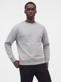 Mand iført en lysegrå crewneck sweatshirt og sorte bukser, stående med den ene hånd på hoften mod en ensfarvet hvid baggrund.