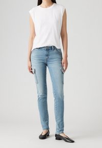 Vaqueros ajustados de mezclilla azul claro con bolsillos laterales, combinados con una blusa sin mangas blanca sencilla y zapatos negros planos, de pie sobre un fondo neutro.