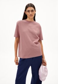 Baumwoll-T-Shirt in Mauve, mit Rundhalsausschnitt und kurzen Ärmeln. Gesticktes Detail auf der Brust. Getragen mit marineblauen Hosen und einem hellrosa Hut in der Hand.