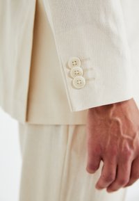Manga de chaqueta de traje de lino beige que presenta tres botones redondos blancos y costuras visibles. Textura suave con un ajuste entallado.