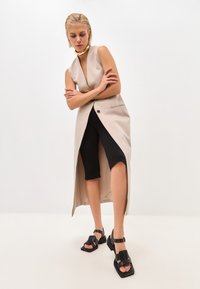 Beige sleeveless vest with a deep V-neck, front button closure, and side slits, paired with black knee-length shorts and chunky black sandals.