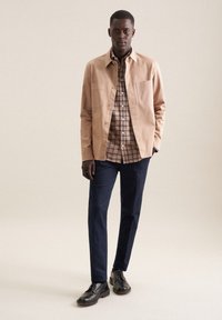 Camicia overshirt beige, camicia a quadri marrone sotto, pantaloni blu navy e scarpe eleganti nere. Texture lisce e vestibilità su misura in evidenza.