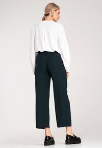 Biała bluzka z długim rękawem i plisą z tyłu, zestawiona z ciemnozielonymi spodniami o szerokich nogawkach. Model nosi czarne buty na platformie. Gładkie tekstury.