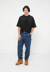 Camiseta negra de gran tamaño con un pequeño gráfico, combinada con pantalones de mezclilla azules y botas marrones, mostrando un ajuste relajado y un estilo casual.